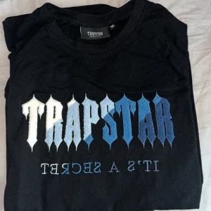 Trapstar svart t-shirt med blått tryck - Svart t-shirt från Trapstar med stort grafiskt tryck i vitt och blått på bröstet. Snygg textdesign och klassisk rund hals. Perfekt för dig som gillar streetwear och vill sticka ut med coola detaljer.
