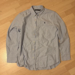 Ljusblå skjorta från Ralph Lauren - Klassisk ljusblå skjorta från Ralph Lauren med button-down krage och broderad logga på bröstet. Skjortan har lång ärm, knappar framtill och är tillverkad i mjuk bomull. Perfekt för en clean och stilren look.