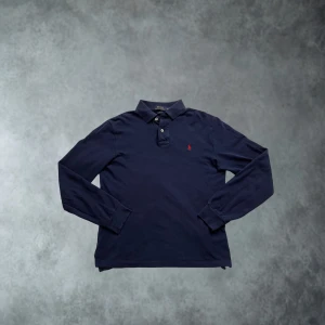 Polo Ralph Lauren tröja/långärmad tröja marinblå o size medium - Klassisk marinblå långärmad pikétröja från Polo Ralph Lauren med röd broderad logga på bröstet. Tröjan har krage, knäppning med två knappar och är tillverkad i mjuk bomull. Perfekt för en stilren och avslappnad look. Tröjan är i ett mycket bra skick och inga skador.                                                      Tveka inte på att skicka ett prisförslag 😀
