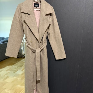 Beige kappa med bälte från ONLY - Säljer en beige kappa från ONLY med breda slag, knytskärp i midjan och dubbelknäppning. Jackan har lång ärm och är tillverkad i ullblandning som ger en mjuk och varm känsla. Perfekt för dig som gillar stilrena och klassiska plagg.