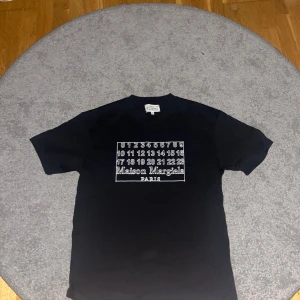 Maison Margiela svart t-shirt - Svart t-shirt från Maison Margiela med ikoniskt tryck framtill med siffror och logga. Klassisk passform, rund hals och korta ärmar. Tillverkad i mjuk bomull för en skön känsla. Perfekt för dig som gillar stilrena och trendiga plagg. 