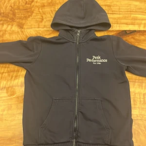 Svart Peak Performance hoodie med dragkedja - Marin blå hoodie från Peak Performance med broderad logga på bröstet. Tröjan har huva, hel dragkedja och två fickor framtill. Mjukt foder på insidan och ribbade muddar vid ärmslut. Perfekt för chill eller träning.