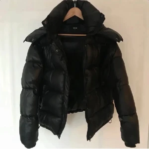 Svart pufferjacka från Bik Bok XS - Svart pufferjacka från Bik Bok i storlek XS. Jackan har en stor huva, dragkedja och knappar framtill samt två sidofickor. Materialet är glansigt och känns lätt, perfekt för kalla dagar. Jackan har en quiltad look och en loose passform.
