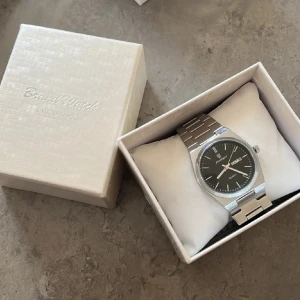 Silverfärgad klocka från Poedagar - Stilren klocka från Poedagar med svart urtavla och dag/datumvisning. Klockan har ett klassiskt armband i silverfärgad metall och minimalistisk design. Perfekt accessoar för dig som gillar en clean look och vill ha koll på tiden.