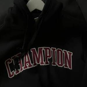 Svart Champion hoodie XS - Svart hoodie från Champion i storlek XS med stor broderad logga i vinrött och vitt över bröstet. Tröjan har huva med snörning, magficka och långa ärmar. Köpt på man-avdelningen så xs kan uppfattas större 