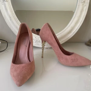 Rosa mockapumps med guldklack - Säljer ett par snygga rosa pumps i mocka från Eastast Blue. Skorna har spetsig tå och en unik, spiralformad guldklack på ca 10 cm som verkligen sticker ut. Perfekta för dig som vill ha något extra! Supersnygga till både jeans och klänning.