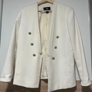 Vit kavaj med silvriga knappar H&M - Stilren vit kavaj från H&M med dubbelknäppning och dekorativa silvriga knappar framtill och vid ärmslut. Rak passform och helfodrad insida. Perfekt för dig som vill ha en clean och modern look.