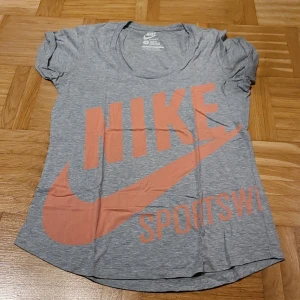 Grå Nike Sportswear t-shirt loose fit - Snygg grå t-shirt från Nike Sportswear med stor orange logga och text framtill. Modellen är loose fit med korta ärmar och rund hals. Perfekt för dig som gillar sportig och avslappnad stil. Tillverkad i mjuk bomull för skön känsla hela dagen.