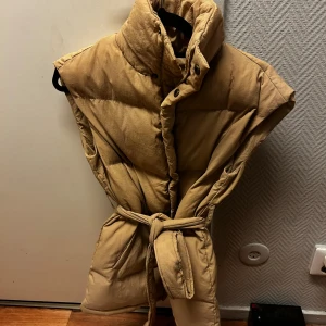 Beige dunväst med bälte och hög krage - Säljer en beige dunväst med hög krage och knytbälte i midjan. Västen har tryckknappar framtill och är quiltad för extra volym. Perfekt att slänga över en hoodie eller tröja när det är kyligt ute. Snygg och enkel att styla till olika outfits.