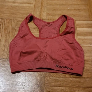 Röd sporttopp från StayInPlace - Säljer en röd sporttopp från StayInPlace med racerback och bred resår nedtill. Toppen är ärmlös och har en tight passform som ger bra stöd vid träning. Materialet är mjukt och stretchigt, perfekt för gymmet eller löpning. Snyggt StayInPlace-tryck på resåren.