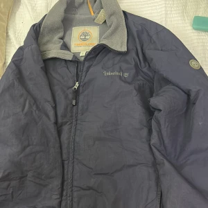 Blå vindjacka Timberland fleecefodrad - Blå vindjacka från Timberland med grått fleecefoder och dragkedja framtill. Jackan har ståkrage, broderad logga på bröstet och patch på ärmen. Perfekt för utomhusaktiviteter och chill stil. Fodret är mjukt och värmande.