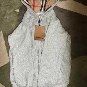 Grå hoodie från Burberry med huva - Snygg grå hoodie från Burberry med klassisk logga och dragkedja framtill. Hoodien har en huva med det ikoniska Burberry-mönstret på insidan, snörning och ribbade muddar. Perfekt för dig som vill ha en stilren och lyxig look.