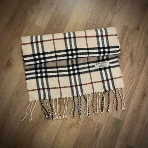 Vintage Burberry rutig halsduk i lammull - Klassisk Burberry halsduk i beige med rutigt mönster i svart, vitt och rött. Gjord av 100% lammull, med fransar i ändarna och ikonisk Burberry-tag. Perfekt accessoar för att lyfta din stil under kyliga dagar.