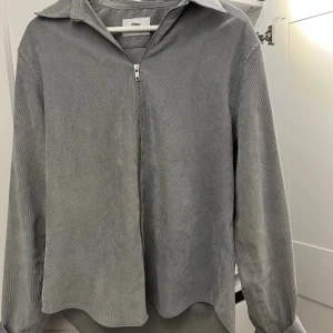 Grå manchester overshirt från Ciszere - Snygg grå overshirt från Ciszere i mjuk manchester. Modellen har klassisk krage, lång ärm och dragkedja framtill. Perfekt att slänga på över en t-shirt för en chill look. Materialet är strukturerat och ger en cool vibe.