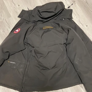 Svart dunjacka från Canada Goose - Svart dunjacka från Canada Goose med huva och klassisk patch på ärmen. Jackan har dragkedja, flera fickor och quiltad insida. Tappat lite färg och använd lite men ser bra ut 
