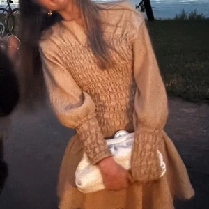 Beige kort klänning med smock - Supersöt beige kort klänning med smockad överdel och ärmar. Klänningen har långa puffärmar och en volangig kjol som ger en romantisk vibe. Perfekt för dig som gillar en feminin och trendig stil.