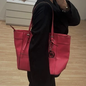 Rosa axelväska från Guess - Snygg illrosa axelväska från Guess med silverfärgad logodetalj och kedja. Väskan har två långa axelremmar, dragkedja upptill och flera fack inuti med mönstrat innerfoder. Tillverkad i stilren design och tydliga sömmar. Sparsamt använd, därav mycket bra skick. Finns endast en liten svart prick på baksidan. 