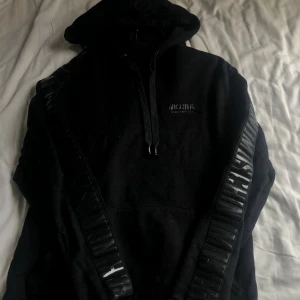 Svart hoodie från Moschino  - Svart hoodie från Moschino Underwear med huva och snörning. Snygga detaljer med Moschino-logga på bröstet och längst ärmarna i blankt svart. Mjuk bomullskvalitet och klassisk passform, perfekt för dig som gillar streetwear och designer.