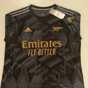 Arsenal svart fotbollströja Adidas  - Snygg Arsenal fotbollströja från Adidas i svart med guldiga detaljer och tryck. Materialet är lätt och ventilerande polyester, perfekt för match eller träning. Oanvänd i perfekt skick. 