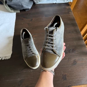 Grå mockasneakers från Lanvin - Snygga sneakers från Lanvin i grå mocka med olivgröna detaljer och tåparti i skinn. Skorna har snörning och vit platt sula. Perfekta för dig som gillar stilrena och unika sneakers med en lyxig touch.