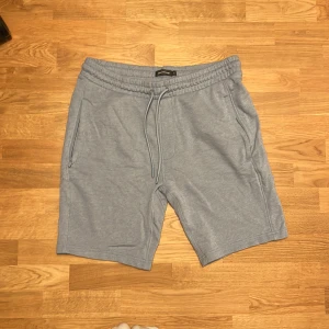 Ljusgrå mjukisshorts från Dressmann - Säljer ett par ljusgrå mjukisshorts från Dressmann i storlek XL. Shortsen har elastisk midja med snörning, två sidofickor och en bakficka. Perfekta för chill dagar eller träning. Tillverkade i mjuk bomullsmix för extra komfort.