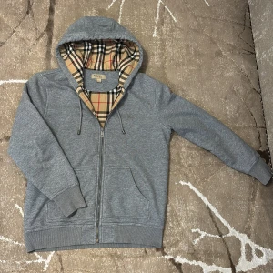 Grå Burberry hoodie med rutig huva - Säljer en grå hoodie från Burberry i storlek L. Tröjan har dragkedja framtill, snörning vid huvan och den klassiska Burberry-rutiga insidan i beige, svart och röd. Tillverkad i 100% bomull, perfekt för dig som gillar stilren design med lyxig touch.