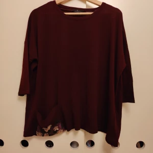 Vinröd loose stickad tröja med paljetter - Vinröd stickad tröja från adl i loose fit med trekvartsärm och rund hals. Nedtill finns en ficka dekorerad med paljetter i beige, svart och lila som ger extra vibe. Perfekt för dig som gillar chill och snygga detaljer.