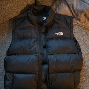 Svart dunväst från The North Face - Säljer en svart dunväst från The North Face med hög krage och diskret logga på bröstet. Västen har en quiltad design och dragkedja framtill. Perfekt för lager-på-lager och streetwear. Materialet är polyester och dun, vilket gör den både varm och lätt.