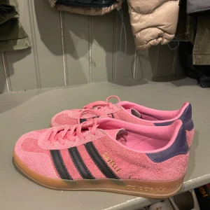 Adidas Gazelle rosa mocka sneakers - Adidas Gazelle sneakers i ljusrosa mocka med klassiska svarta ränder på sidorna och mörkblå detaljer vid hälen. Skorna har snörning och platt gummisula i beige. Perfekt för dig som vill sticka ut med färg och retrostil.