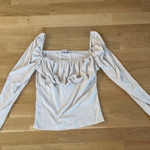 Beige ribbad blus från Bubbleroom - Supersöt beige blus från Bubbleroom i storlek S. Den har ribbat tyg, puffiga långa ärmar och en fyrkantig urringning. Volangdetaljer vid bysten ger en extra touch. Perfekt för dig som gillar romantisk och trendig stil.