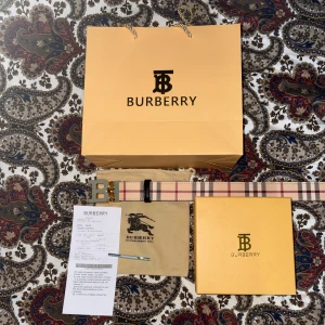 Burberry randigt bälte med guldspänne - Säljer ett snyggt Burberry bälte med klassiskt randigt mönster i beige, rött och svart. Bältet har ett stilrent guldspänne med Burberrys B-logga. Perfekt accessoar för att lyfta din outfit med lyxig känsla och ikonisk design.