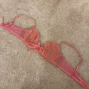 Rosa spetsbralette med rosetter - Säljer en snygg rosa bralette med blommig spets och flera små rosetter. Modellen har justerbara axelband och hakknäppning bak. Spetsen är genomskinlig och ger en feminin vibe, perfekt för dig som gillar detaljer och en lite lyxigare känsla.