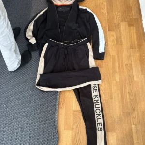 Svarta mjukisbyxor Moose Knuckles M - Svarta mjukisbyxor från Moose Knuckles i storlek M med beige sidopaneler och logotyp längs benet. Byxorna har snörning i midjan och är gjorda i mjukt bomullsmaterial. Snygg sportig look med kontrastdetaljer och bekväm passform.