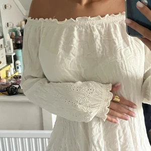 Vit offshoulder blus med broderade ärmar - Säljer en vit offshoulder blus med långa ärmar och broderade detaljer. Blusen har en rynkad kant upptill och är tillverkad i ett lätt, krinklat bomullstyg. Ärmarna har fina broderier och hålmönster som ger en extra touch. Perfekt för dig som gillar romantisk stil.