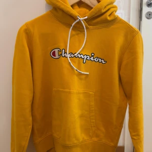 Gul hoodie från Champion XS - Gul hoodie från Champion i storlek XS med broderad logga på bröstet. Tröjan har huva med vit snörning, känguruficka och långa ärmar. Perfekt för dig som gillar streetwear och vill sticka ut med en färgglad look.