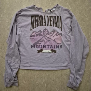 Tröja - Ljuslila långärmad croppad tröja från H&M med tryck av Sierra Nevada Mountains i lila och svart. Rund hals med liten slits framtill, mjukt bomullsmaterial och avslappnad passform. Perfekt för dig som gillar coola prints och chill vibe.