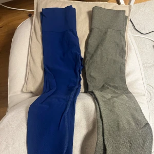 Blå och grå leggings med medel hög midja - Två par leggings, ett i klarblått och ett i grått. Båda har bred linning och är tillverkade i stretchigt syntetmaterial som sitter tight mot kroppen. Perfekta för träning eller chill hemma. Snygg passform och enkel design utan mönster eller detaljer.