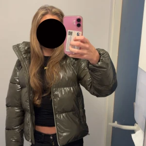 Mörkgrön pufferjacka med huva - Säljer en mörkgrön pufferjacka med glansig finish och huva. Endast använd 1 gång. Jackan har dragkedja framtill och är quiltad för extra värme. Perfekt för kalla dagar. Slutsåld på hemsidan