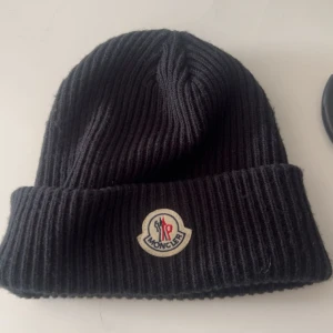 Svart ribbad mössa från Moncler - Svart ribbstickad mössa från Moncler med klassisk uppvikt kant och ikonisk Moncler-logga framtill. Perfekt för kalla dagar och ger en clean look. Materialet är mjukt och värmande, och den svarta färgen gör den enkel att matcha med allt.