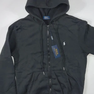 Svart hoodie från Polo Ralph Lauren - Svart hoodie från Polo Ralph Lauren med klassisk vit logga på bröstet. Tröjan har huva med snörning, dragkedja framtill och en stor ficka på magen. Perfekt för dig som gillar stilrena och bekväma plagg med premiumkänsla.