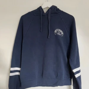 Marinblå hoodie från Gina Tricot - En marinblå hoodie från Gina Tricot med vita ränder på ärmarna och tryck på bröstet. Tröjan har huva med snörning och är tillverkad i mjuk bomullsmix. Perfekt för allmän användning och enkel att matcha med annat 