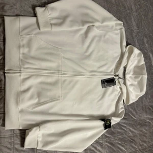 Vit hoodie från Stone Island - Säljer en clean vit hoodie från Stone Island med klassisk logopatch på vänster ärm. Tröjan har huva, dragkedja framtill och två stora fickor. Materialet är mjuk bomull och passformen är normal. Perfekt för dig som gillar stilrena streetwear-plagg. Tröjan har QR Scan och priset går även att diskutera vid snabb affär.