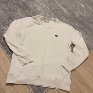 Vit sweatshirt från Lacoste - Vit sweatshirt från Lacoste i klassisk passform med rund hals och ribbade muddar. Tröjan har det ikoniska gröna krokodilmärket broderat på bröstet. Perfekt för dig som gillar stilren och enkel design.