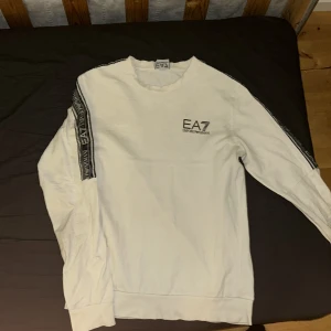 Vit EA7 sweatshirt med logga - Snygg vit sweatshirt från EA7 Emporio Armani med svarta loggband längs ärmarna och tryckt logga på bröstet. Tröjan har rund halsringning och långa ärmar, perfekt för en clean och sportig look.