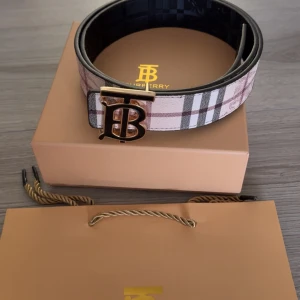 Rutig Burberry bälte med guldspänne - Snyggt bälte från Burberry med klassiskt rutigt mönster i beige, svart, vitt och rosa toner. Bältet har ett stilrent guldigt 'TB'-spänne framtill och är tillverkat i skinn. Perfekt accessoar för att lyfta din outfit med en lyxig touch.