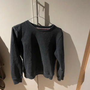 Mörkgrå sweatshirt Tommy Hilfiger - Mörkgrå sweatshirt från Tommy Hilfiger med klassisk rund hals och diskret logga på bröstet. Tröjan har ribbade muddar vid ärmslut och nederkant samt långärmad passform. Perfekt för chill dagar och enkel att matcha med jeans eller joggers.