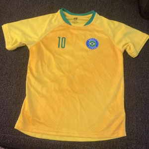 Gul Brasil fotbollströja - Snygg gul Brasilien fotbollströja med gröna detaljer och nummer 10 på både fram- och baksidan. Tröjan har korta ärmar, rund hals med grön kant och Brasilien-emblem på bröstet. Tillverkad i lätt och skönt polyester-material.
