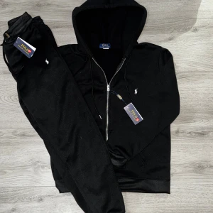 Svart Polo Ralph Lauren Tracksuit - Svart hoodie från Polo Ralph Lauren med dragkedja och huva. Klassisk vit logobrodering på bröstet, snörning i huvan och mjukt bomullsmaterial. Perfekt för chill dagar och streetwear-stil. Snygg och enkel design med ribbade muddar och fickor framtill. Taggar sitter kvar och helt ny ete kvalite