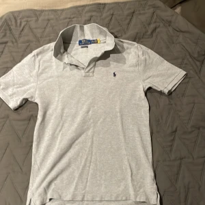Grå pikétröja från Polo Ralph Lauren - Snygg grå pikétröja från Polo Ralph Lauren med klassisk passform och korta ärmar. Tröjan har en stilren krage och den ikoniska lilla broderade loggan på bröstet. Perfekt för en chill och clean look, enkel att matcha med jeans eller shorts. Storleken funkar till både xs och s. Ny pris ca 1200