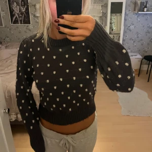 Mörkgrå prickig stickad tröja - Supermysig mörkgrå stickad tröja med vita prickar och ribbade muddar. Modellen är croppad med rund hals och har långa ärmar med lite puff. Perfekt för dig som gillar en söt och chill stil. Passar dig som vill ha något mjukt och varmt till hösten eller vintern.